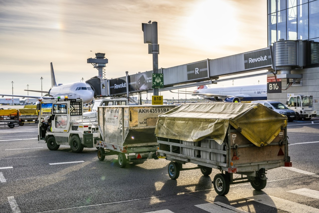 A small vehicle with cargo trailers, an aeroplane at the gate in the background.