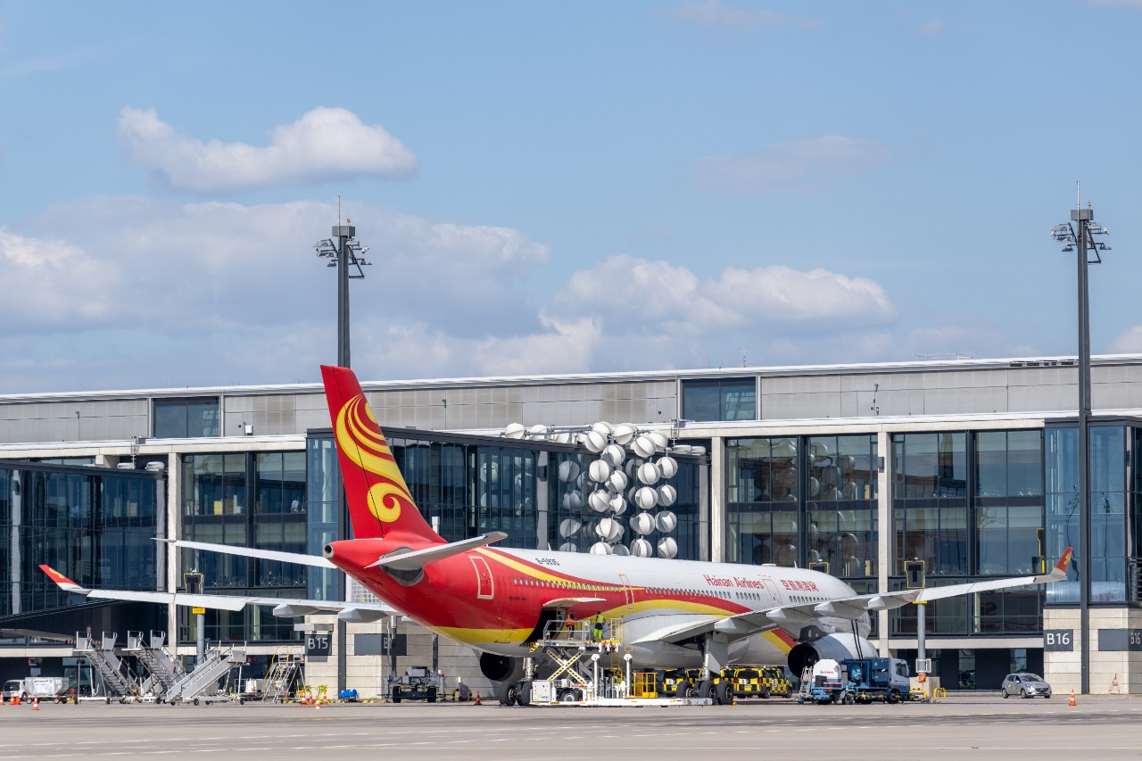 The Airbus A 330 of Hainan Airlines stands at its handling position at BER. In the background you can see the glass façade of Terminal 1 and an art installation decorating the jetway between the terminal building and the aircraft.