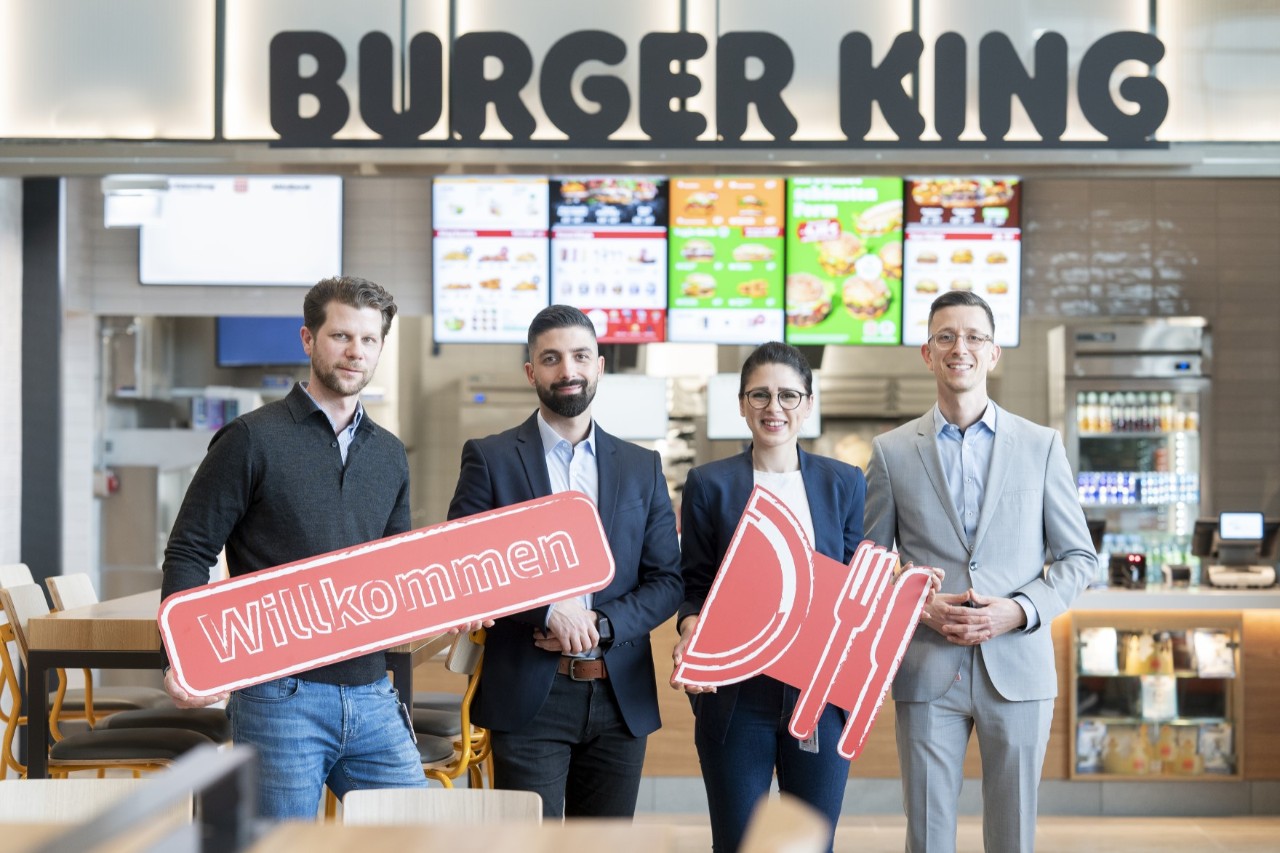Three men and a woman are standing in front of a Burger King branch; they are holding up red signs: one of the signs says "Welcome", the other is in the shape of cutlery and a plate