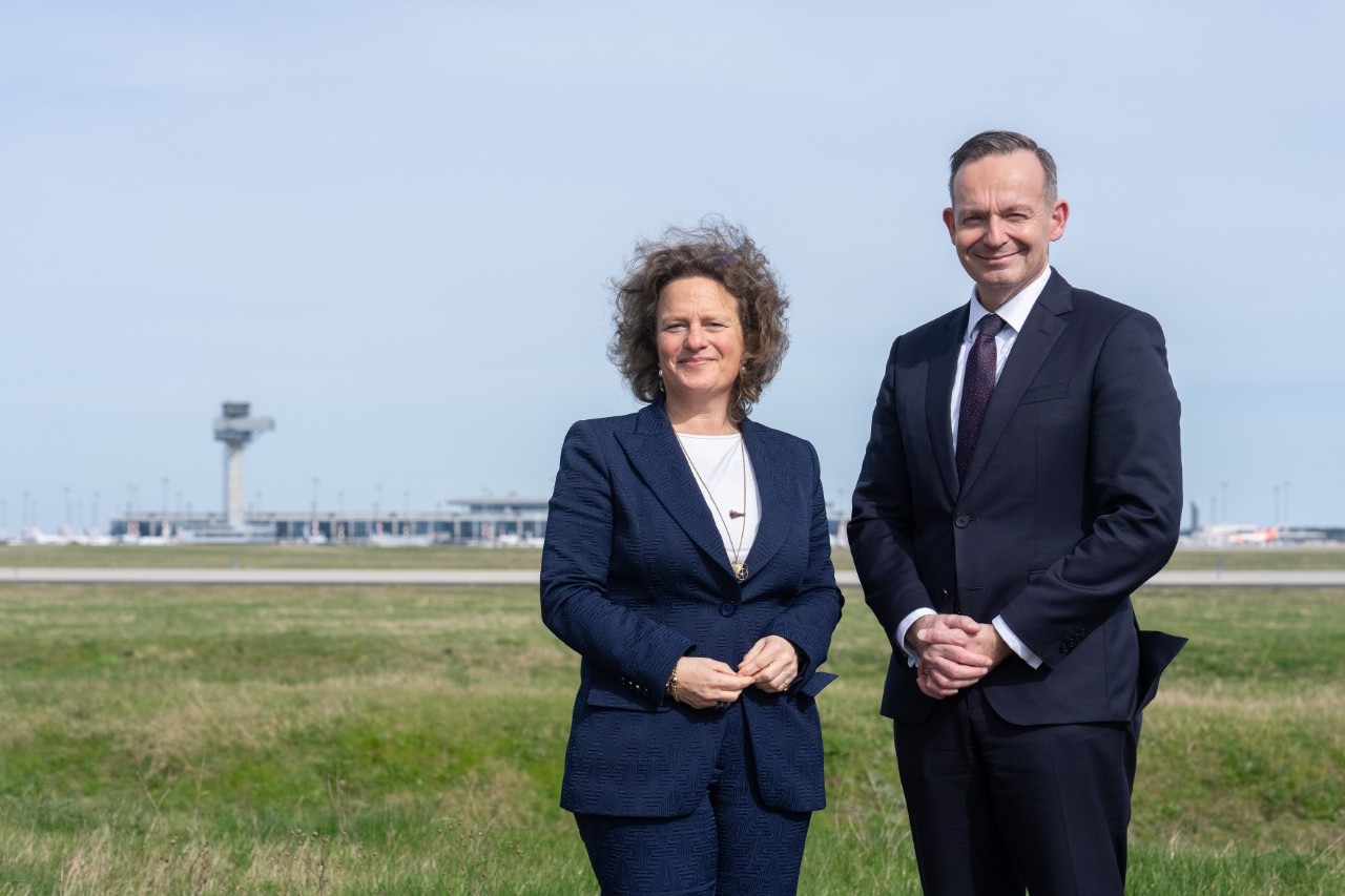 A woman and a man stand in front of a wide meadow. The outline of BER airport can be seen in the background