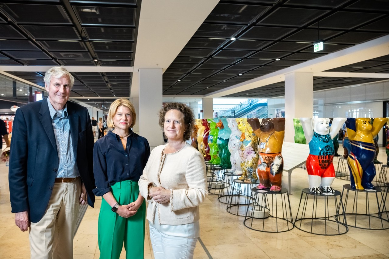 Three people stand in a terminal and smile at the camera. Mini Buddy Bears stand on pedestals in the right background.