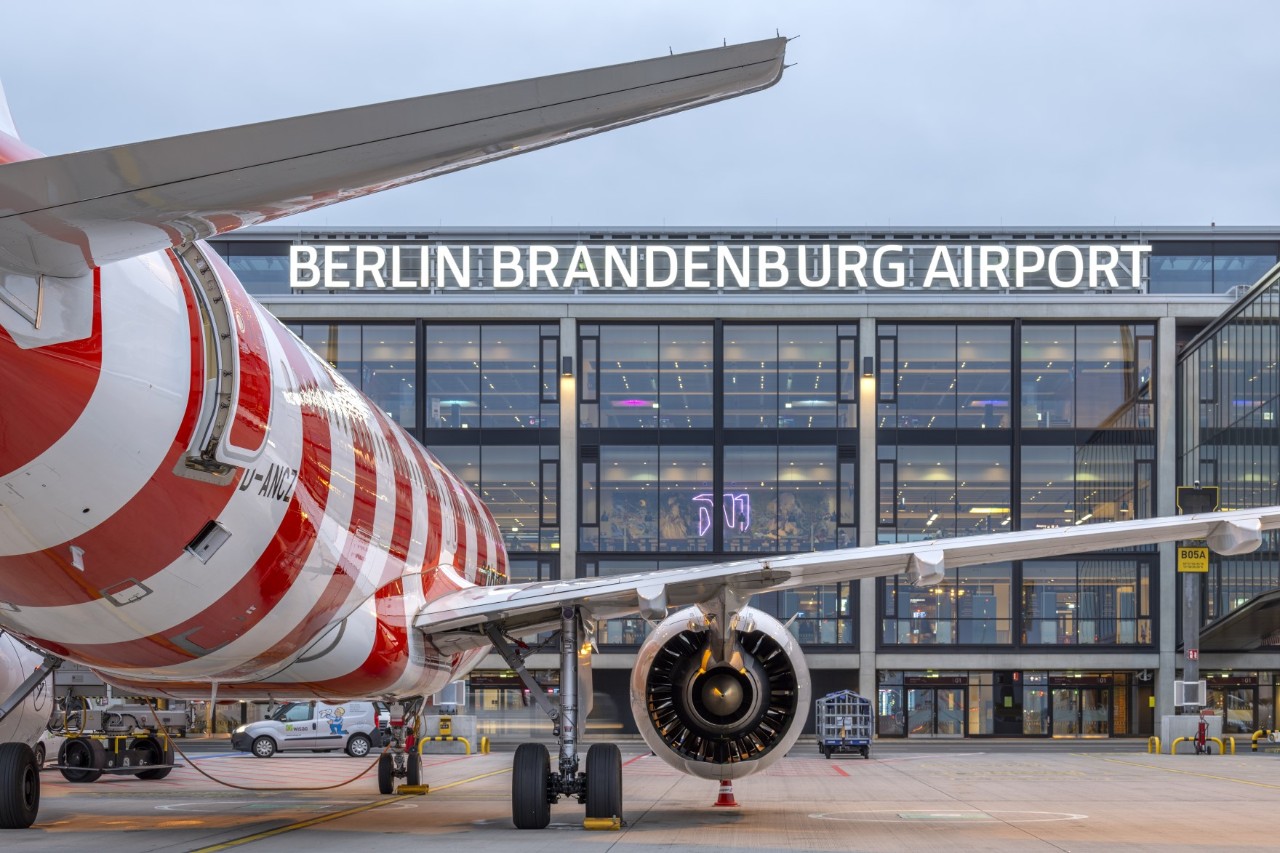 A red and white striped aircraft is parked on the apron in front of the BER terminal. ‘Berlin Brandenburg Airport’ is written on the glass façade.