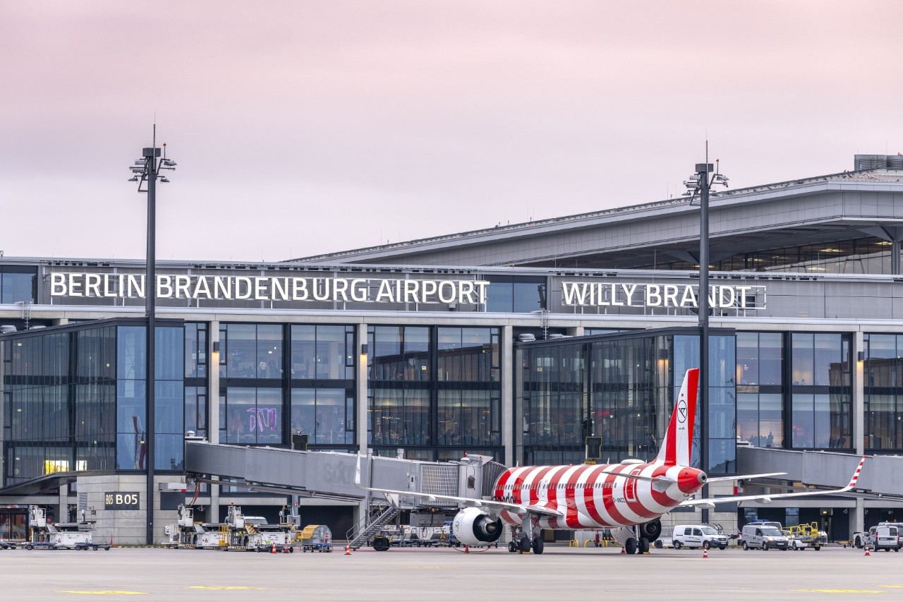 A red and white striped Condor aircraft is parked in a ground handling position directly in front of the BER terminal. A passenger boarding bridge is docked to the aircraft.