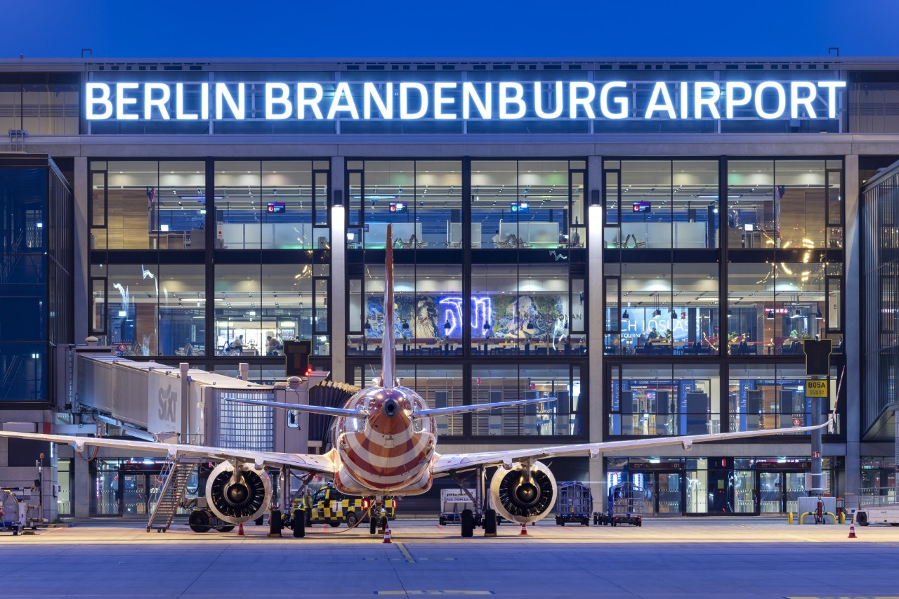 A red and white striped aircraft stands in a ground handling position directly in front of the BER terminal, which is illuminated at night. The illuminated lettering ‘Berlin Brandenburg Airport’ is attached to the façade of the terminal.