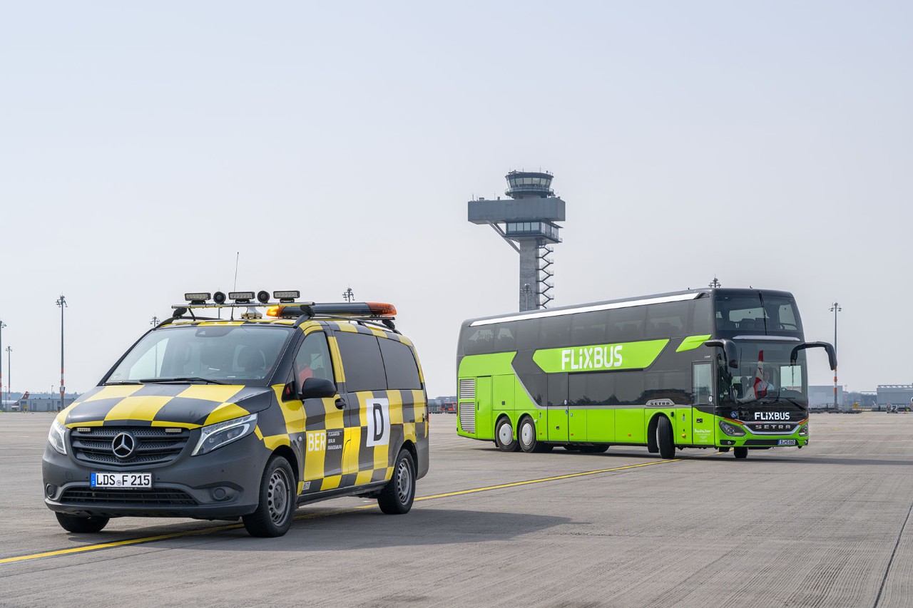 A follow-me vehicle is parked on the apron. In the background, a green long-distance coach is driving past.