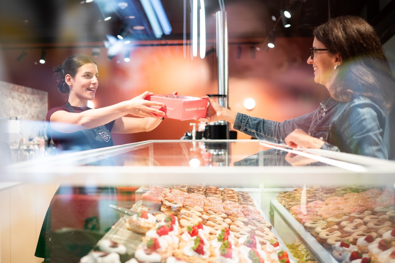 A shop assistant hands a box to a customer over a counter full of doughnuts.