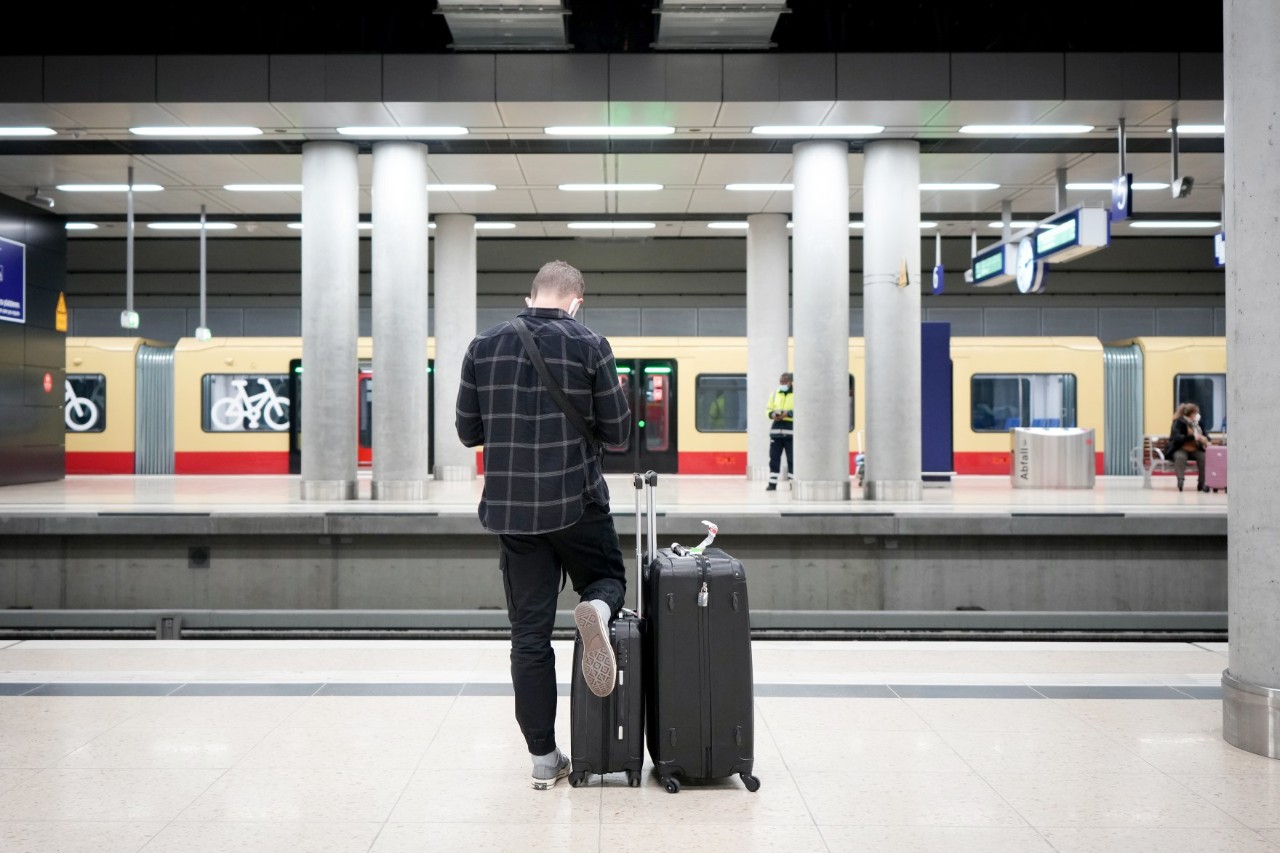 A passenger with a suitcase is standing on a platform at BER airport station. An S-Bahn train is standing on another platform in the background.