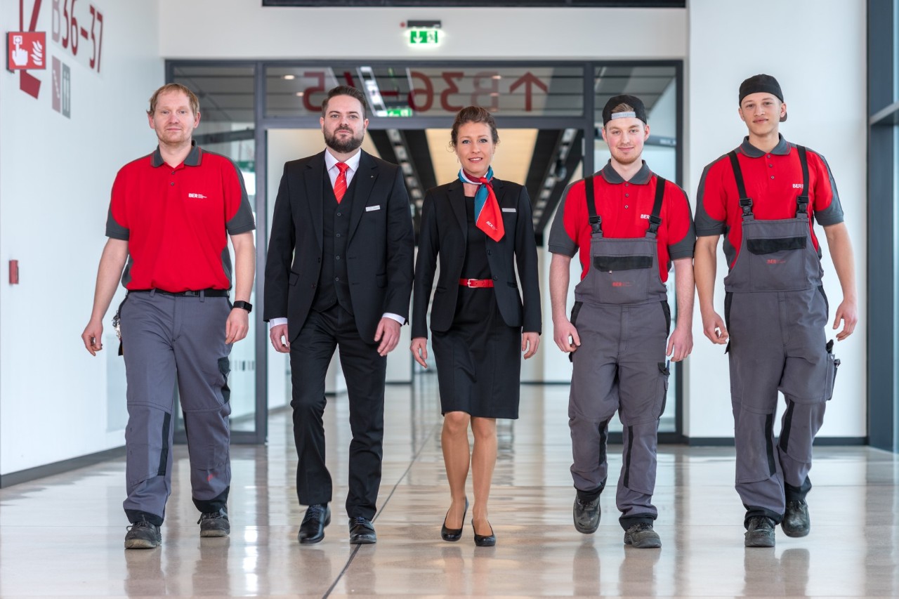 Five people in different work uniforms walk towards the camera, smiling.