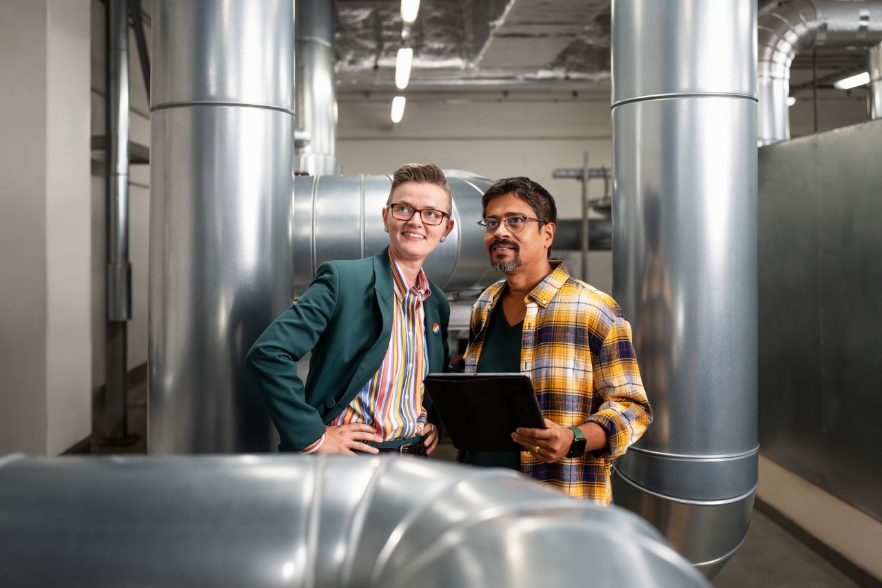 Two employees in a large room with various pipes running through it