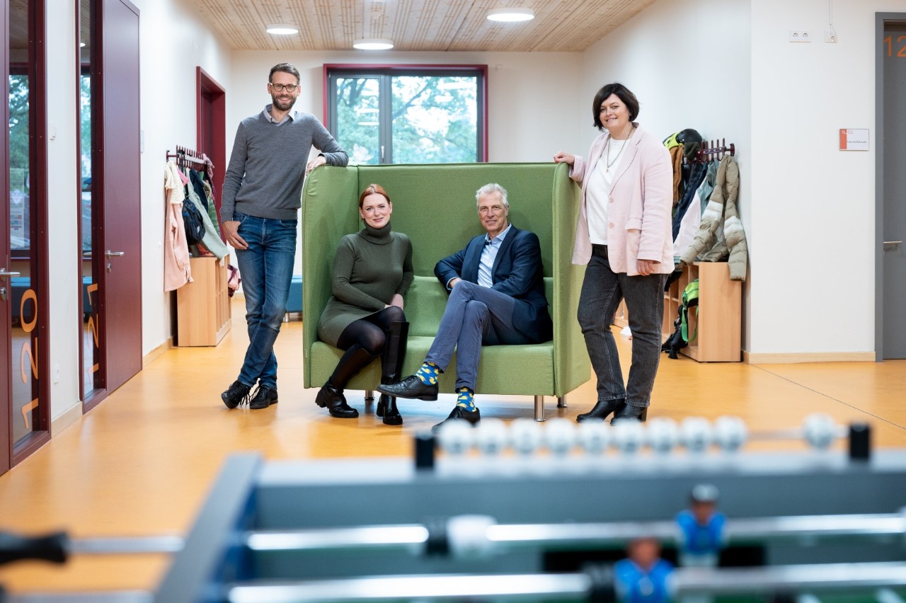 Four adults are smiling as they pose in a school corridor. Two of them are sitting on a sofa with high sides in the middle of the corridor.