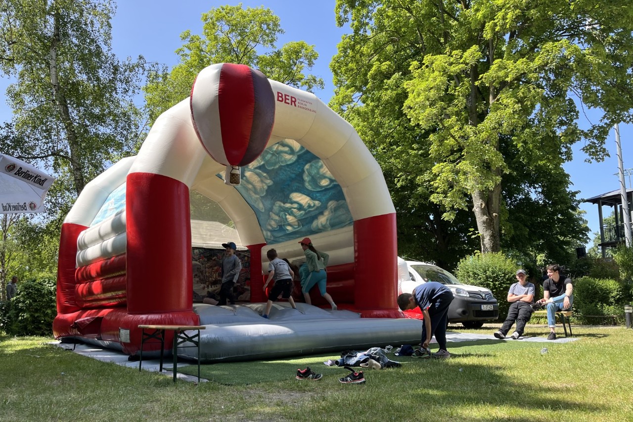 Children are jumping on a bouncy castle with the BER logo, which has been set up on a meadow under a blue sky.